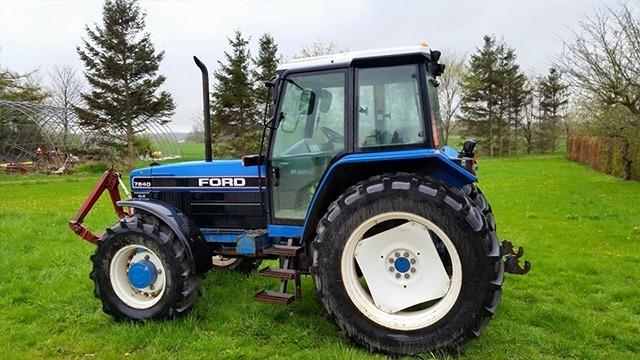 En blå Ford traktor parkeret på en grøn græsmark med træer i baggrunden.