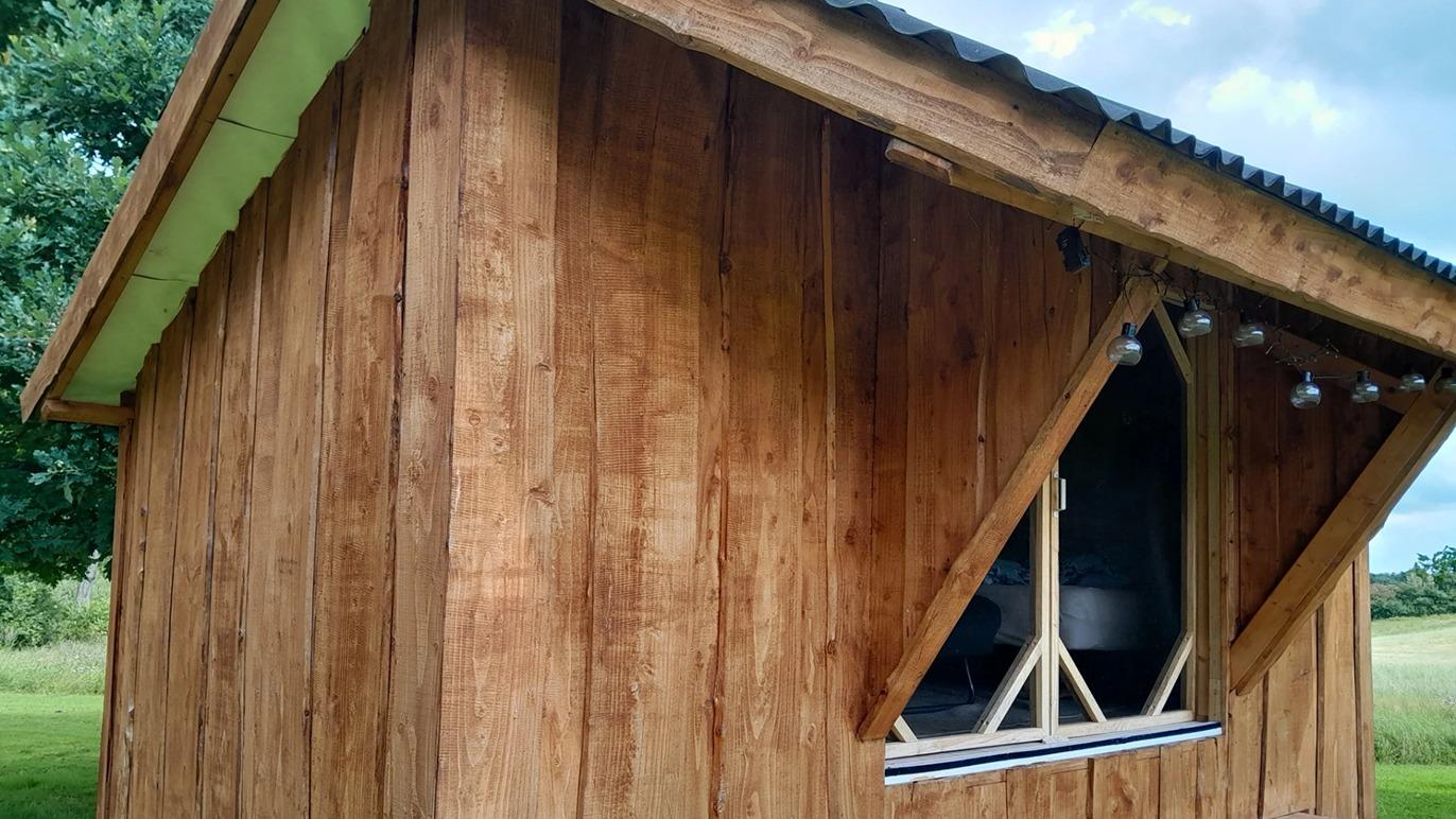 Et lille træhus med en skrå tag og vindue i en trekantet form, omgivet af grønne marker.