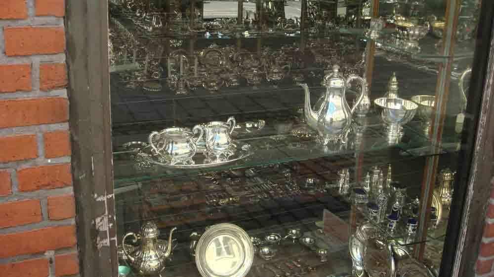 Vitrineskab med sølvbestik, herunder tepotter, sukkerkander og tallerkener, udstillet i en butik.