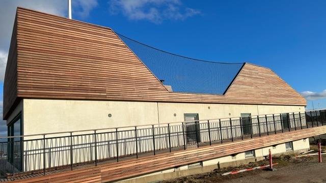 Moderne bygning med skrå tag og træbeklædning, omgivet af en terrasse og blå himmel i baggrunden.