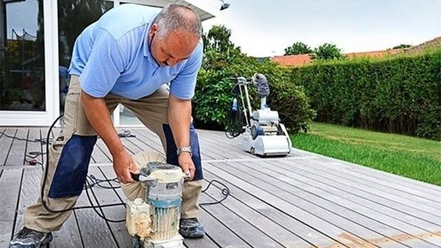En mand arbejder med en maskine på en træterrasse i en have. Græsset er grønt i baggrunden.