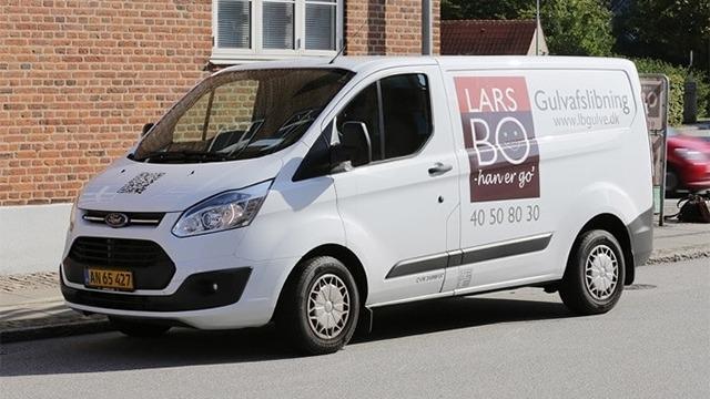 Hvid varevogn med teksten "LARS BO Gulvbelægning" parkeret på en gade.