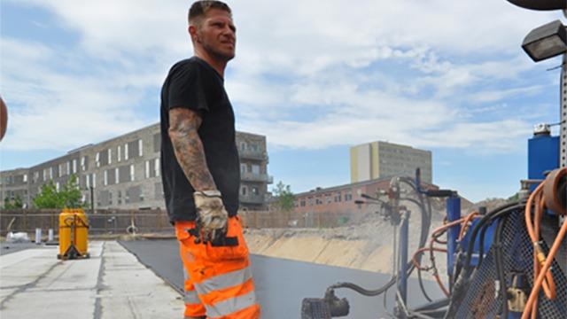En mand i orange arbejdstøj står ved en maskine på en byggeplads under en blå himmel.