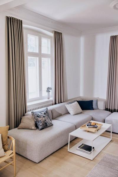 Moderne stue med en grå sofa, et hvidt sofabord og store vinduer med gardiner.