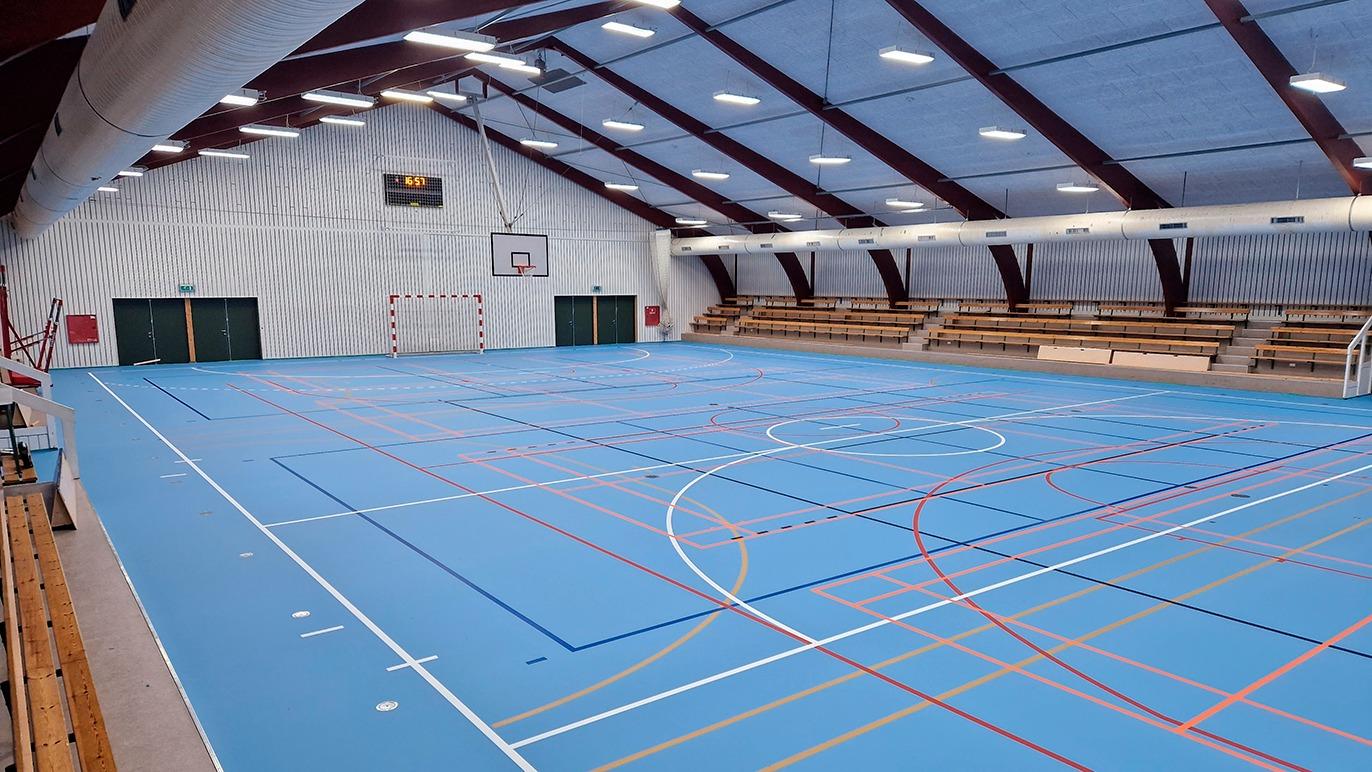 Indendørs sportshal med blåt gulv, basketballkurv og tribuner til tilskuere.