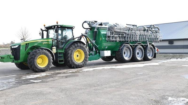 En grøn traktor trækker en stor gyllevogn på en grusvej ved en landbrugsbygning.