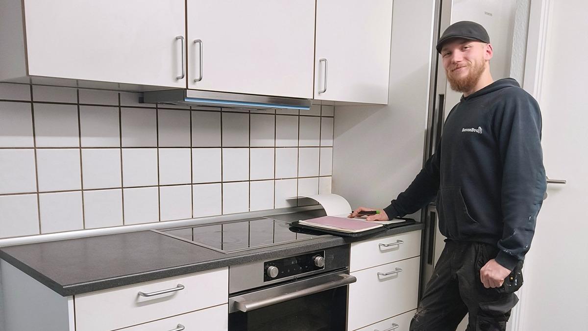 En mand står ved et moderne køkken med en komfur og en vask, iført sort tøj og kasket.