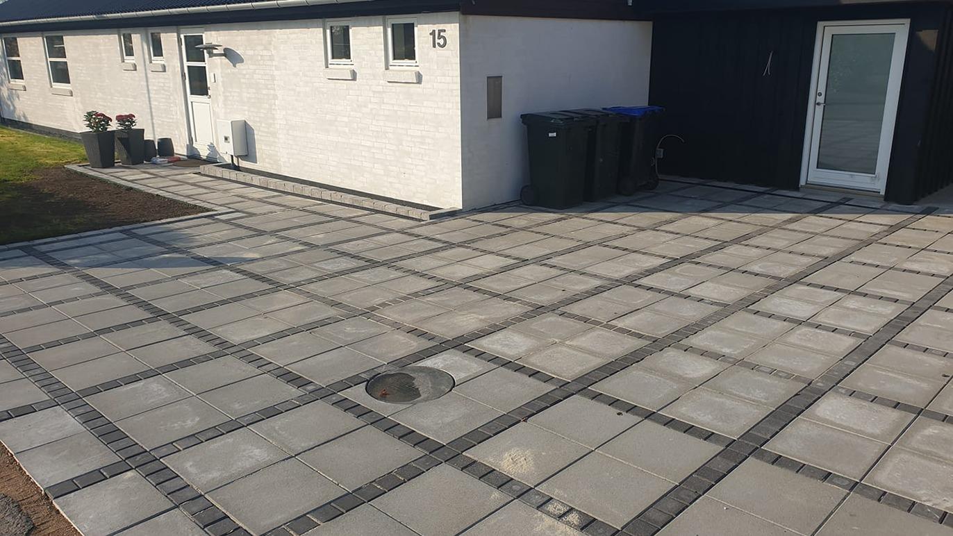 En moderne indkørsel med grå fliser og affaldscontainere ved en hvid murstensbygning.