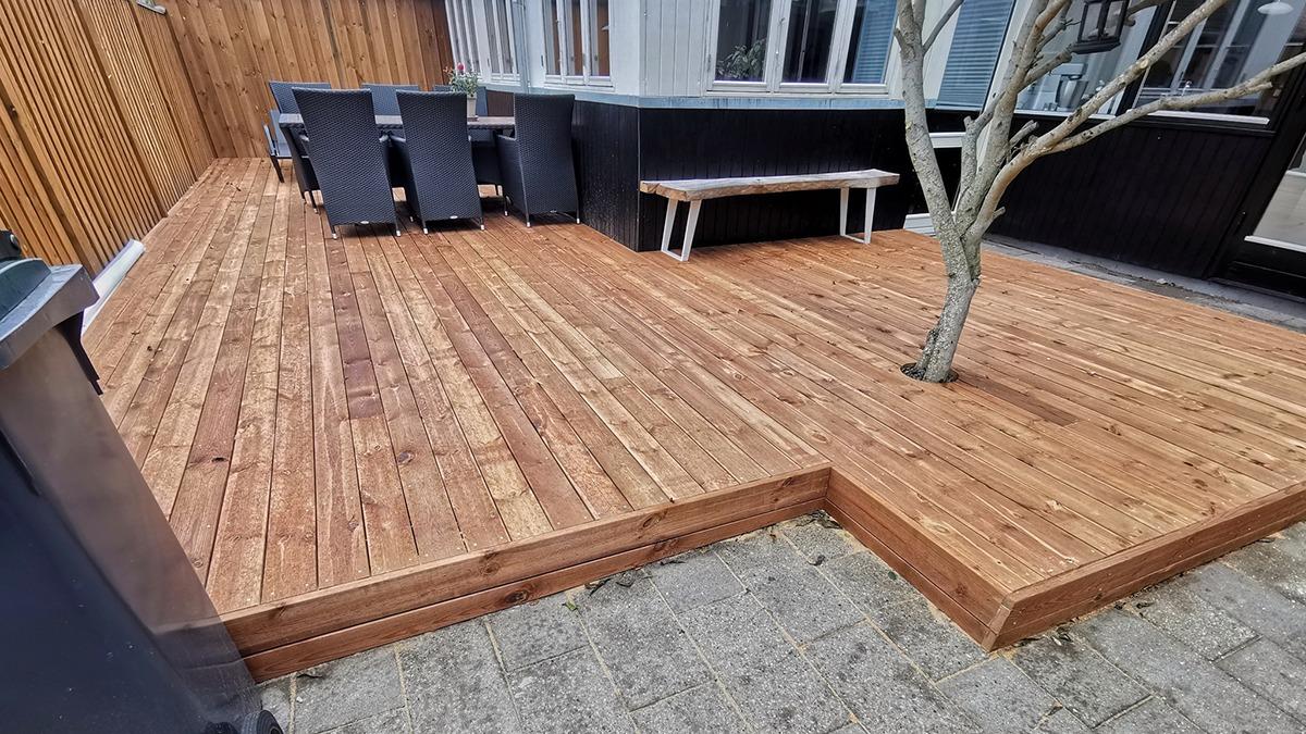 Ny træterrasse med en træbænk og en parasol, omgivet af en træhegn og et hus i baggrunden.