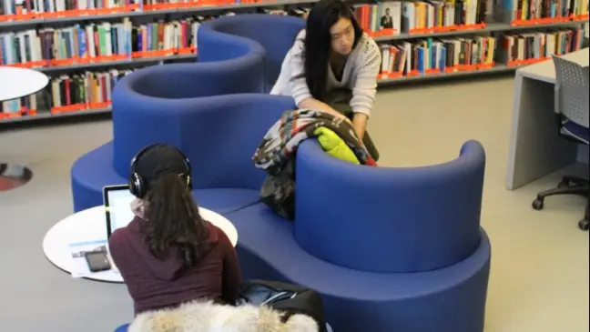 To personer sidder i et bibliotek, en på en blå sofa med hovedtelefoner og en computer, den anden ordner bøger.