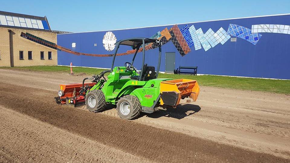 En grøn minitraktor arbejder på en grusvej med en redskabssammensætning bagpå.