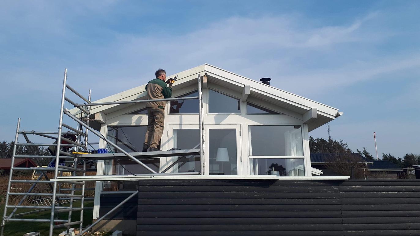 En håndværker arbejder på taget af et hus, mens han står på en stige under en blå himmel.