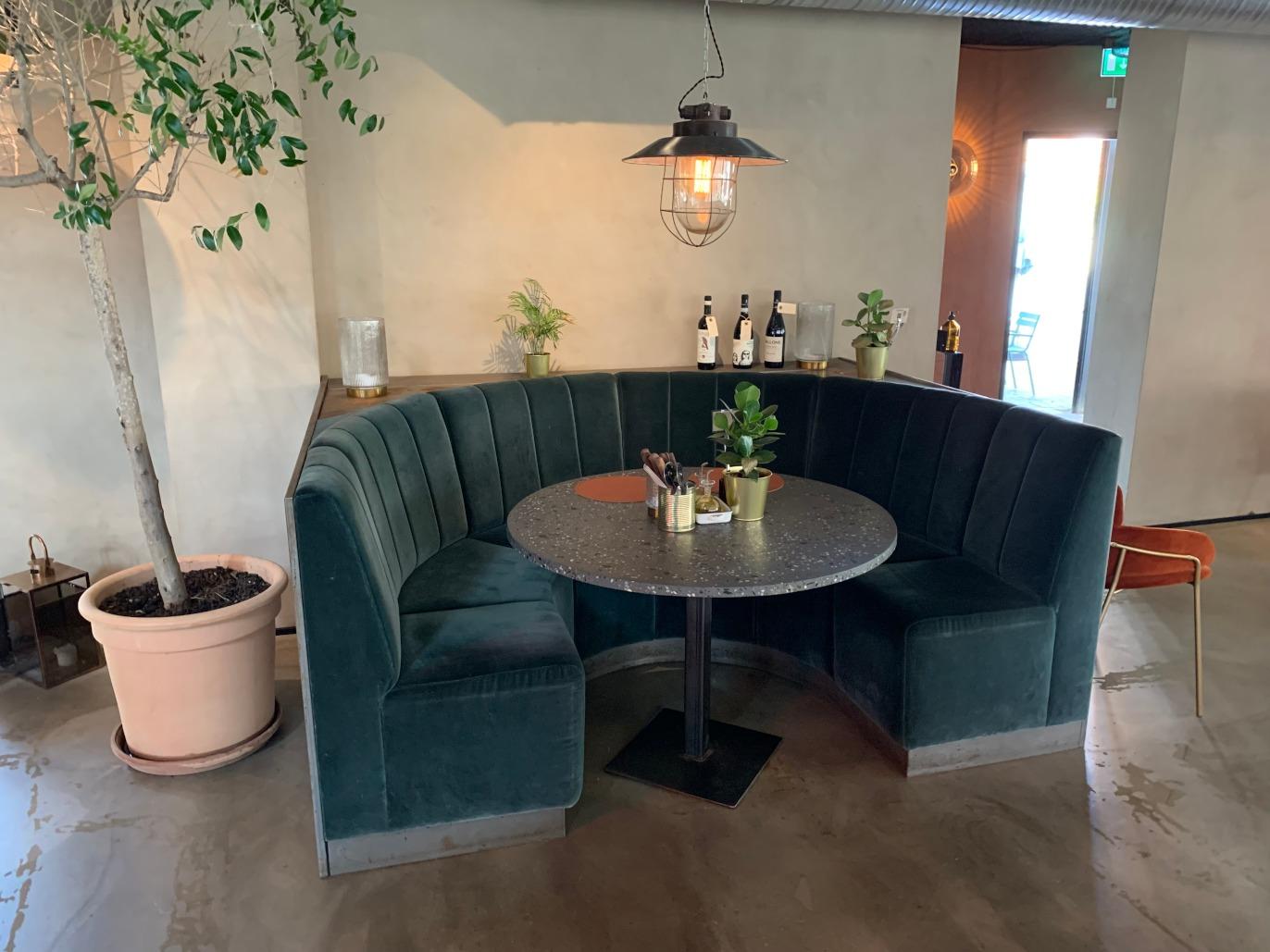 Hyggeligt hjørne med en rund bordplade, grønne sofaer og planter i en moderne restaurant.