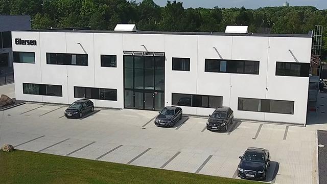 Moderne kontorbygning med glasindgang og flere parkerede biler foran. Grønne træer i baggrunden.