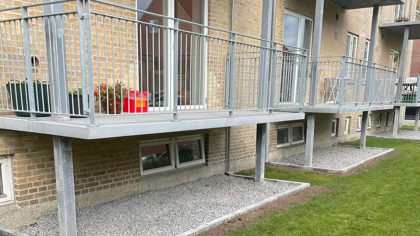 Balkoner på en bygning med grå gelænder, planter og grusbelægning foran.