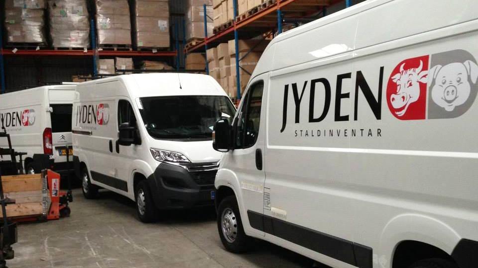 Tre hvide varevogne med JYDEN-logo parkeret i et lagerområde med kasser i baggrunden.