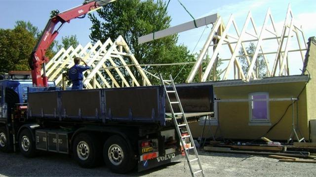 Byggeri af et hus med en kran, der løfter tagkonstruktion, mens en arbejder overvåger processen.