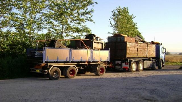 Lastbil med trailer fyldt med genstande og trælast, parkeret ved en grusvej omkranset af træer.
