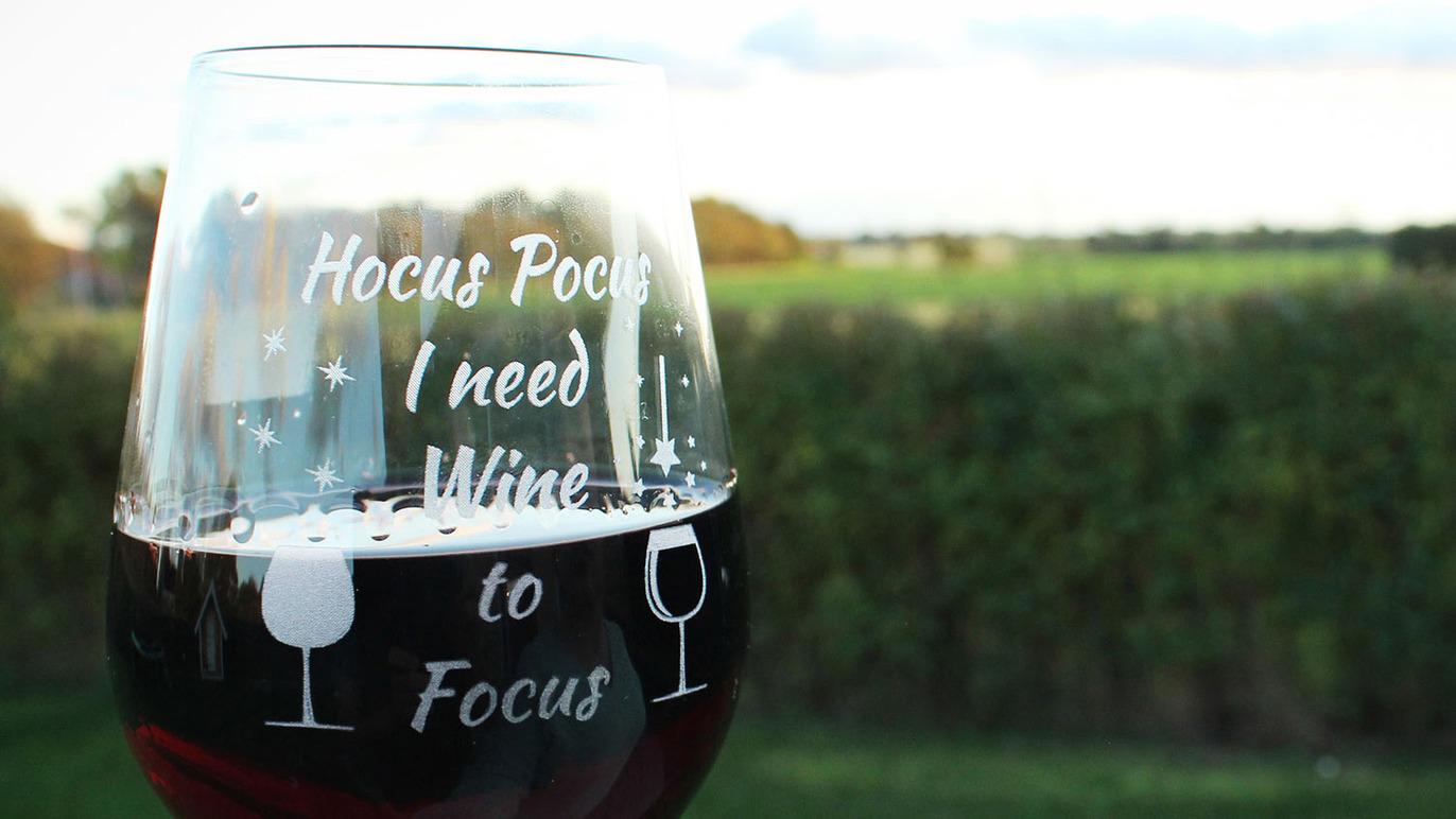 Et vinglas med teksten "Hocus Pocus, I need Wine to Focus" og en grøn mark i baggrunden.