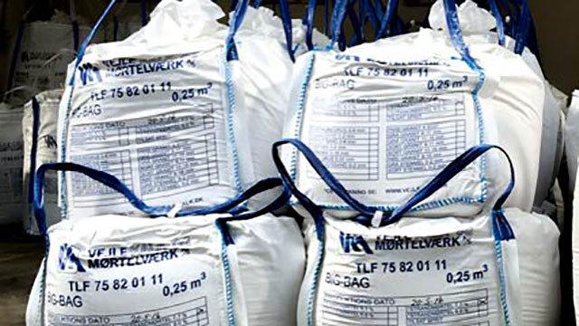 Store sække med byggematerialer stablet i en lagerhal, mærket med produktinformation og vægt.