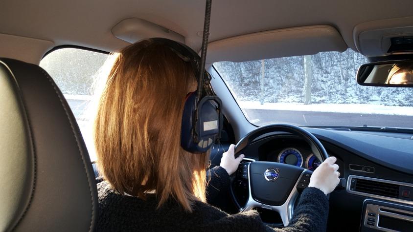 En person med kort hår, der kører bil, iført hovedtelefoner, med sneklædte træer i baggrunden.