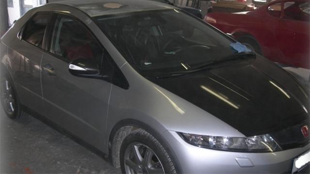 En sølvgrå bil med sort motorhjelm parkeret i en garage med andre biler i baggrunden.
