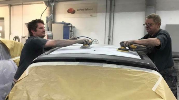 To mænd arbejder på at slibe taget af en bil i et autoværksted. Bilen er dækket med beskyttende plast.