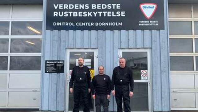 Tre mænd står foran Dinitrol Center Bornholm, der tilbyder rustbeskyttelse. Skiltet viser "Verdens Bedste Rustbeskyttelse".