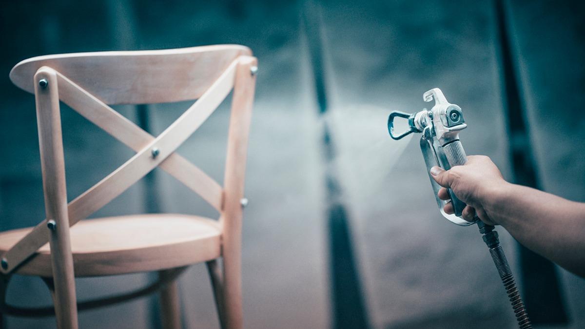 En hånd holder en malerpistol, der sprayer farve på en træstol i et malerværksted.