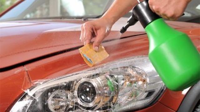 En person rengør en bils forrude med en sprayflaske og en svamp. Bilen er orange og står i et lyst miljø.