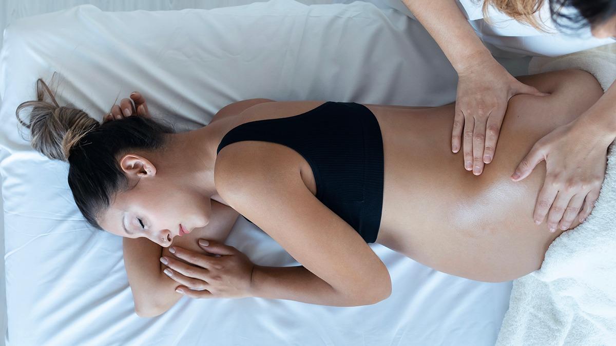 Gravid kvinde får massage på en hvid seng, mens hun ligger på siden med lukkede øjne.