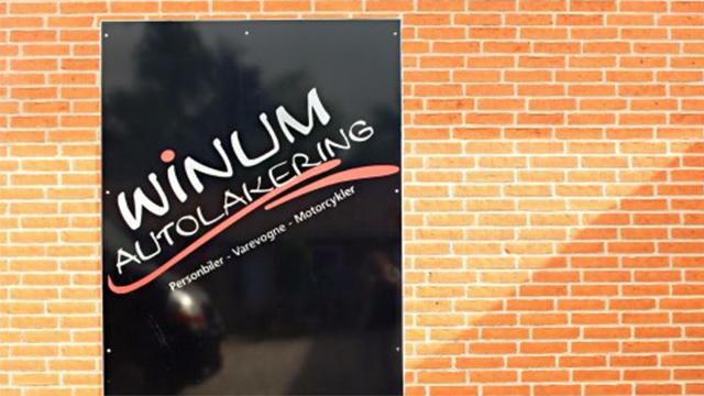Skilt med teksten "WINUM AUTOLAKERING" på en murstensvæg, der reklamerer for autolakeringstjenester.
