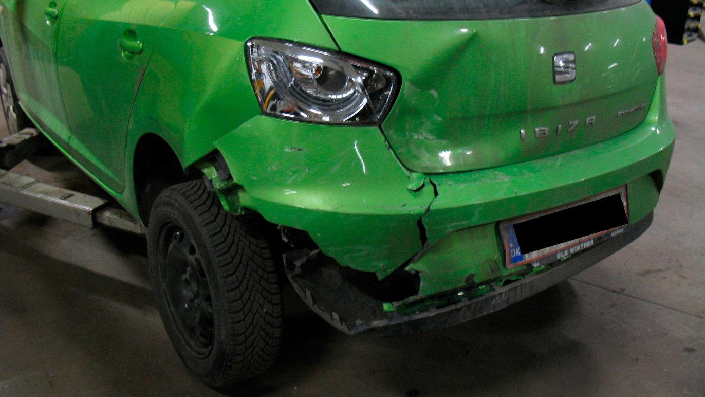 Skadet grøn bil med buler og ridser på bagenden, parkeret i en garage.