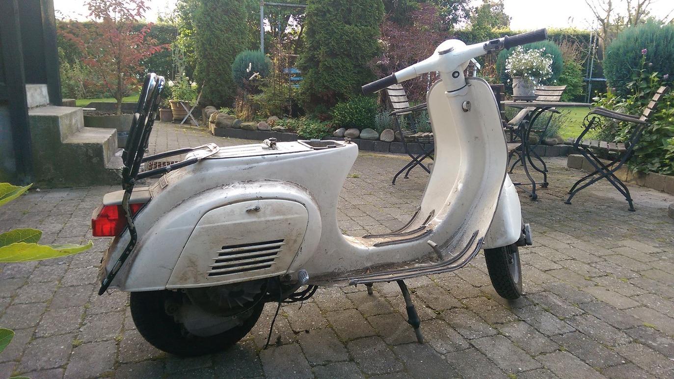 En hvid scooter parkeret på en flisebelagt gårdhave med planter og havemøbler i baggrunden.