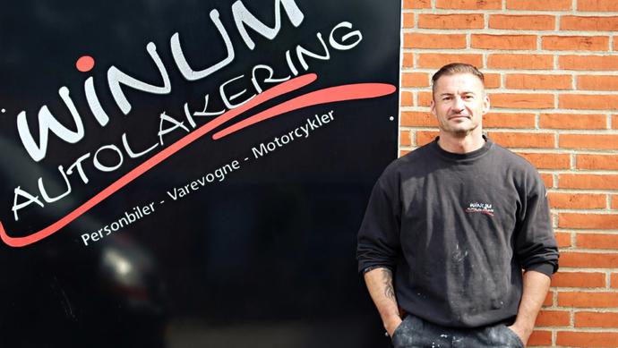 En mand står foran en væg med logoet for Winum Autolakering, der tilbyder bil- og motorcykelmalerarbejde.