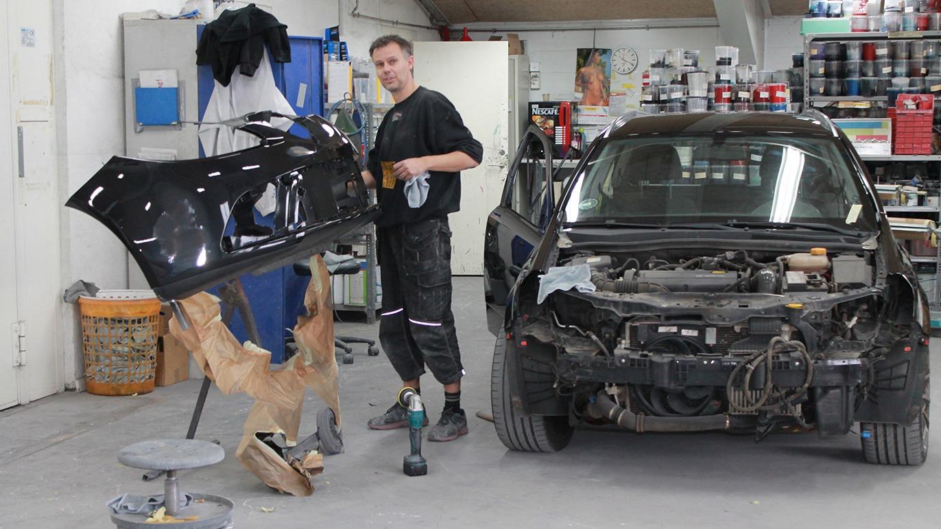 En mekaniker arbejder på en bil i et værksted, mens han holder en bilforkofanger i hånden.