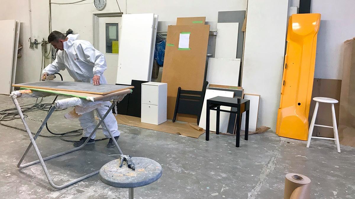 En person i hvidt arbejdstøj maler en plade i et værksted med forskellige materialer og møbler i baggrunden.