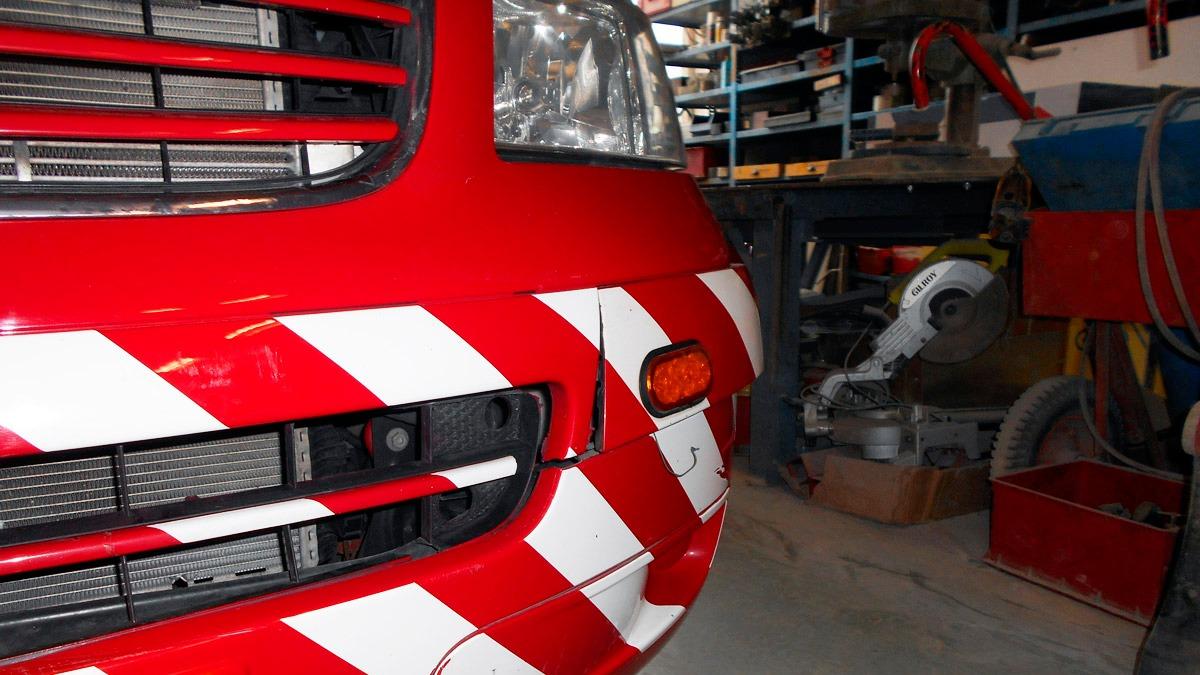 Rød bil med hvide og røde striber i en garage med værktøj og maskiner i baggrunden.