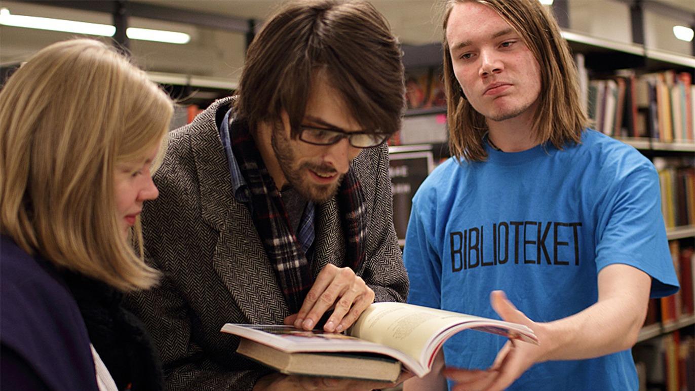 Tre unge mennesker ser på en bog i et bibliotek, mens de diskuterer indholdet.