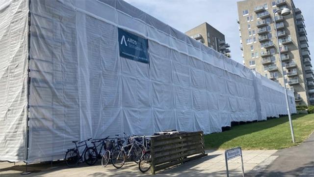 Bygning dækket af hvidt stillads med cykler parkeret foran og grøn græsplæne i forgrunden.