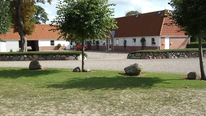 En gård med rødt tag, grusvej og store sten i forgrunden, omgivet af træer og grøn græsplæne.