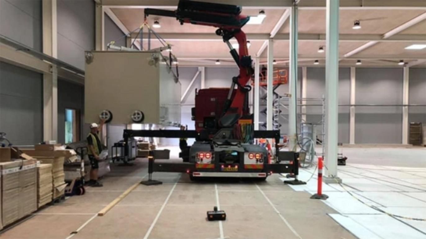 En kran arbejder på en byggeplads med en stor container og en arbejder i sikkerhedshjelm i baggrunden.