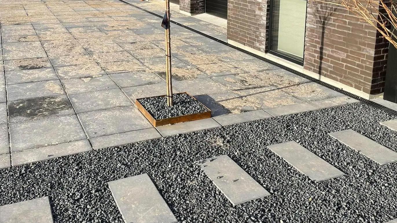 Flisebelagt gårdhave med grus og en lille træplante i en kasse.