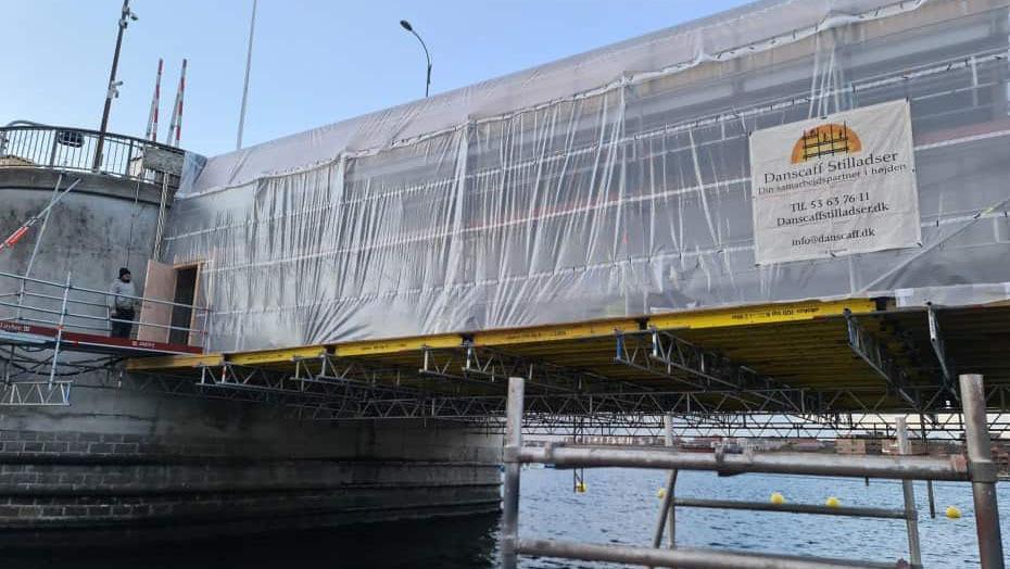 Brobyggeri med stillads og plastikoverdækning ved vandet, med et skilt om arbejdet.