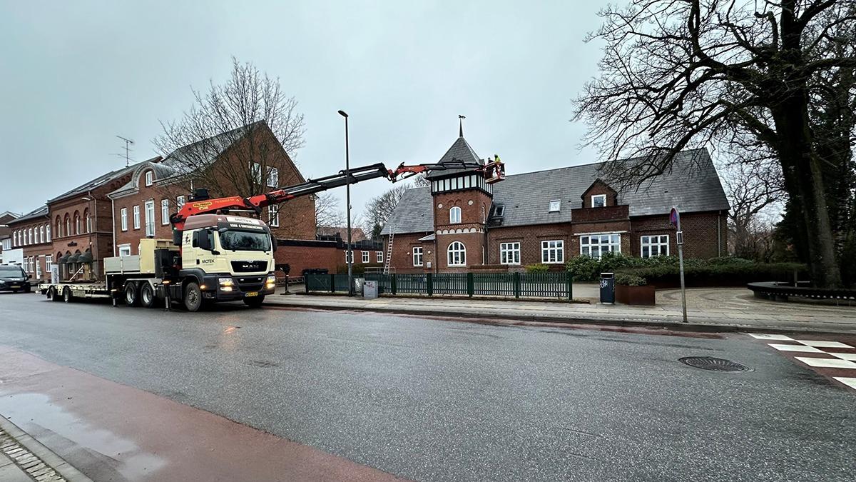 Lastbil med kran parkeret på en vej foran en bygning med tagvindue og træer i baggrunden.