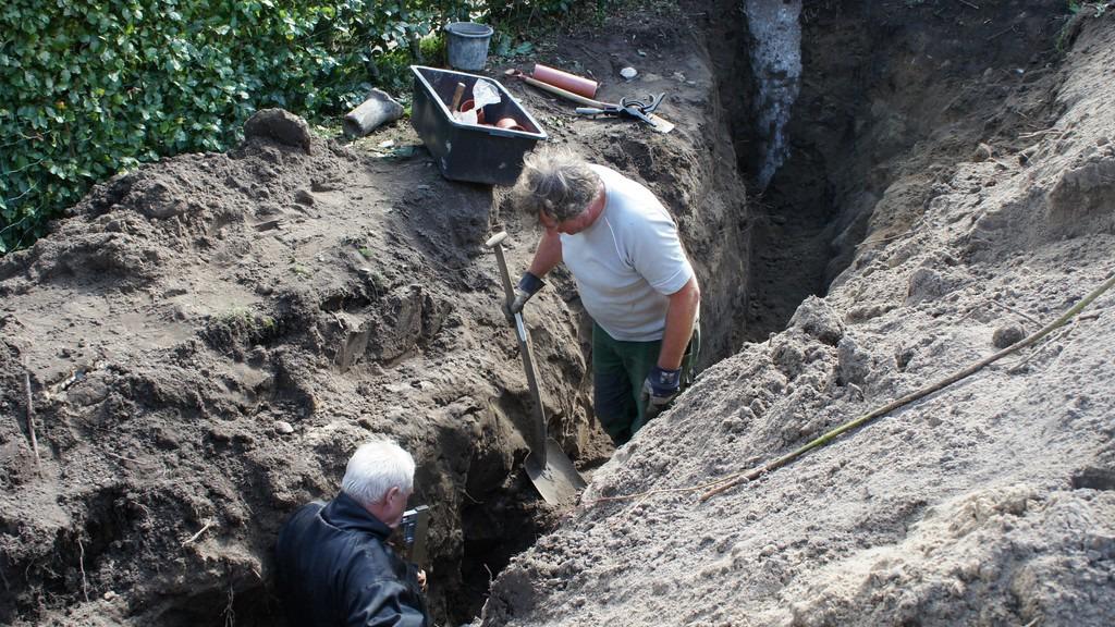 To personer graver i en dyb grøft fyldt med jord og værktøj, omgivet af grønne planter.