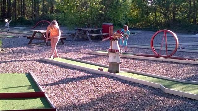 Børn spiller minigolf på en udendørs bane med grønne områder og legepladsudstyr i baggrunden.