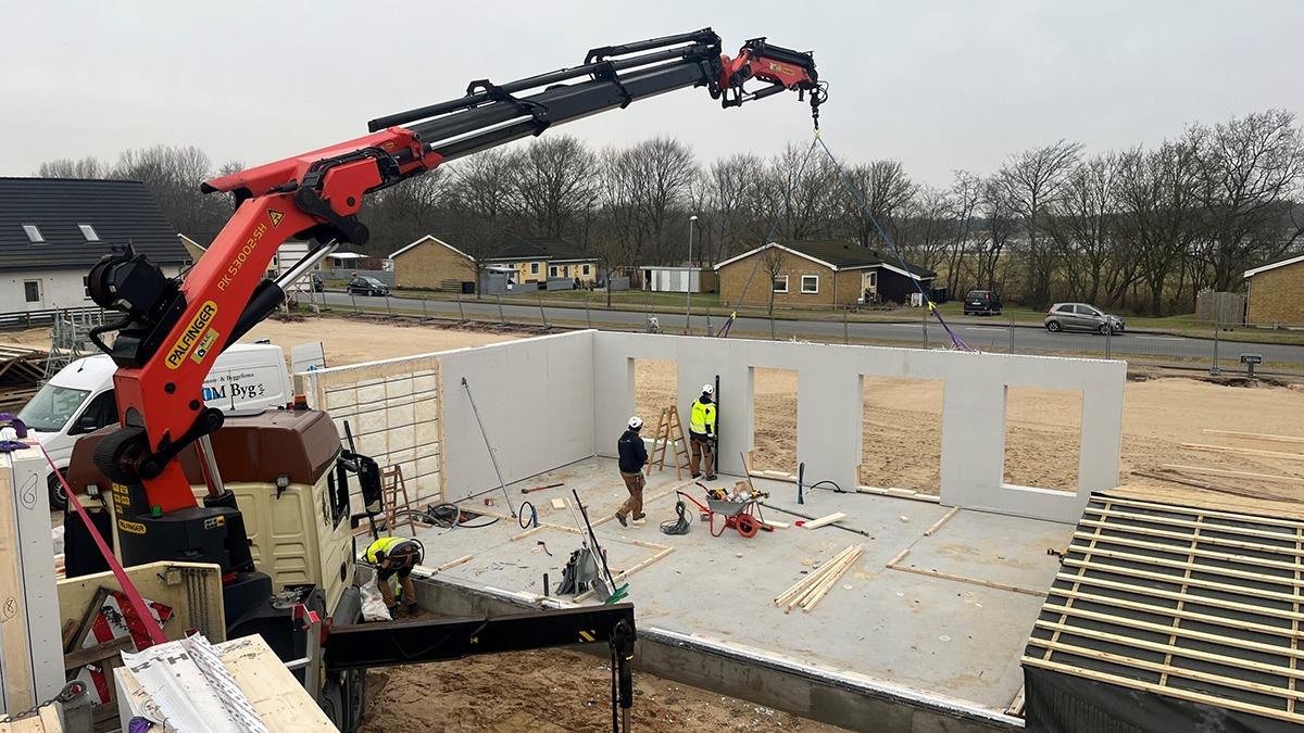 Byggeplads med kran, der løfter vægge, mens arbejdere arbejder på fundamentet til et hus.