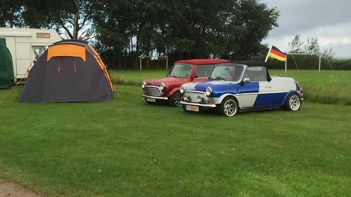 To biler, en rød og en blå, parkeret ved en campingplads med et telt i baggrunden og en tysk flag.