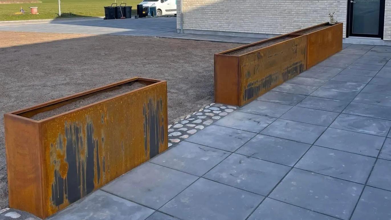 To store plantekasser i cortenstål på en flisebelagt gård ved en moderne bygning.